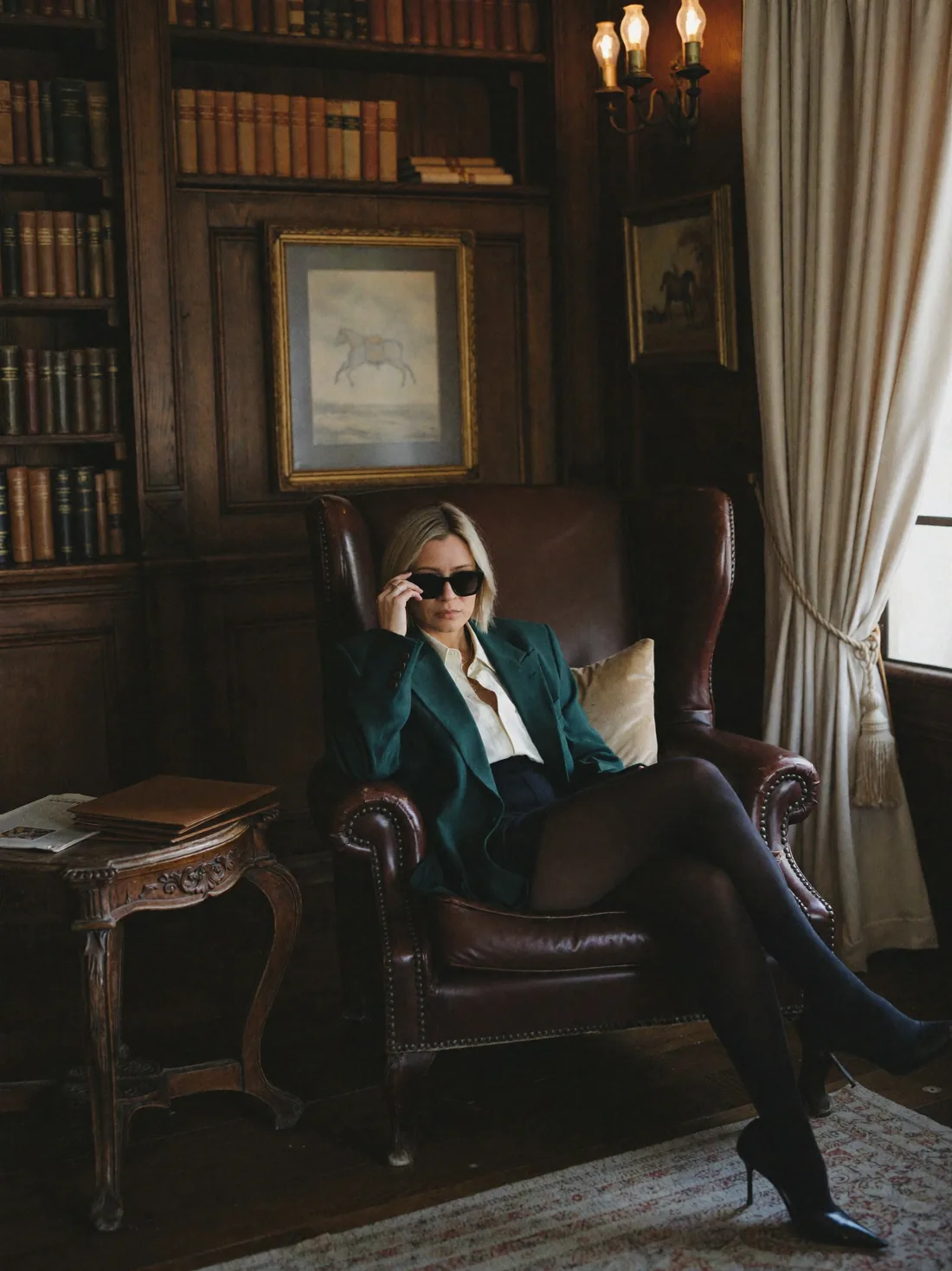 Amanda in a green blazer and sunglasses, sitting in a leather armchair in a library